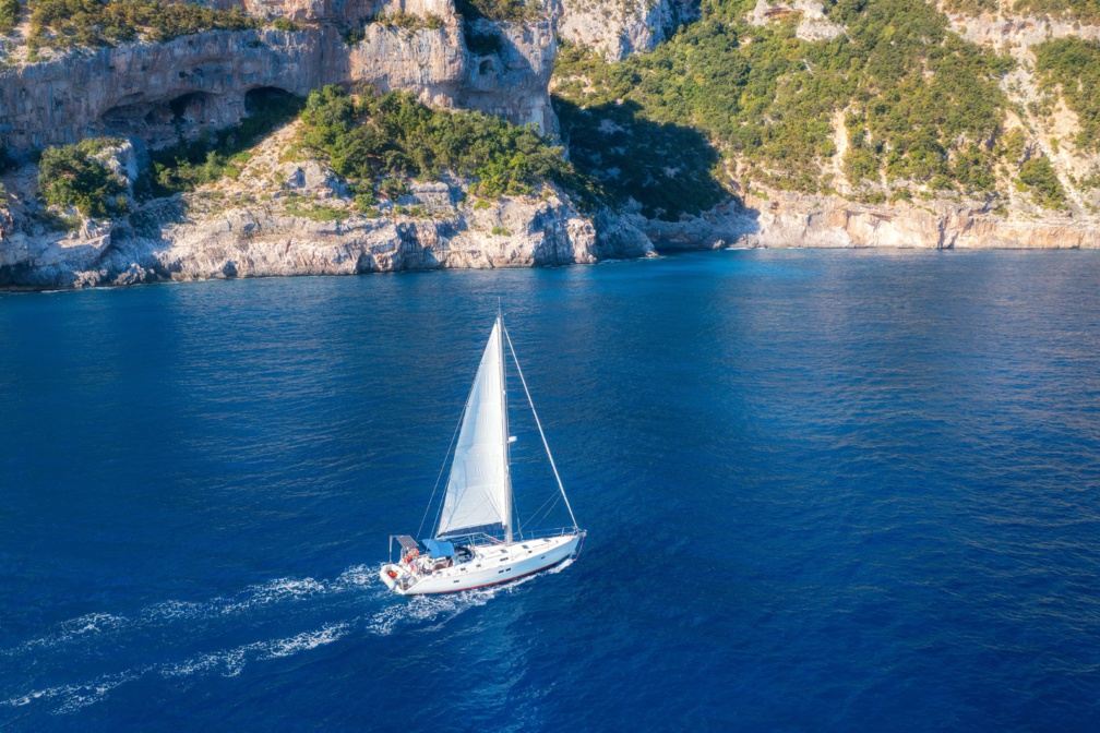 Vue aérienne d'un voilier flottant et d'une côte rocheuse lors d'une journée d'été ensoleillée. Vue aérienne d'un yacht, de montagnes, d'arbres et d'eau bleue. Yachting en Sardaigne, Italie. Paysage marin tropical avec bateau dans l'océan © den-belitsky - stock.adobe.com